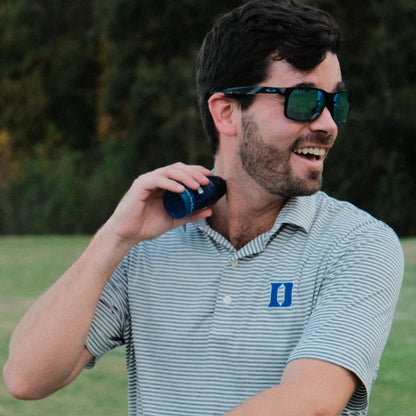 Man wearing sunglasses and a striped polo shirt holding a Pacto sunscreen outdoors.