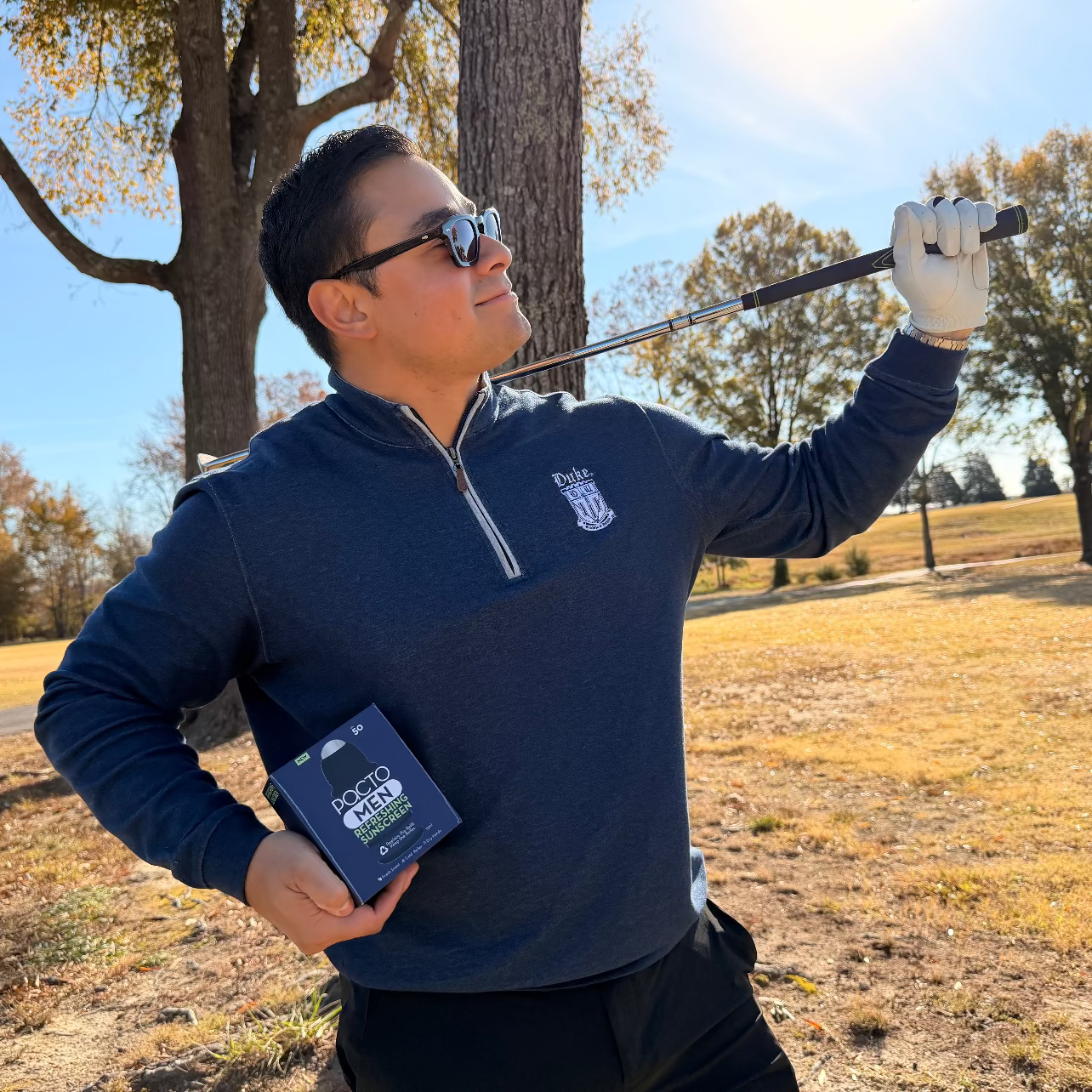 Man holding a golf club and a device in a field with trees in the background